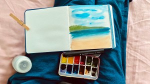 Watercolor book with a landscape painted on it, paint palette, and travel brush in my lap. There’s a clothespin holding open the left side of the paper in the book. 