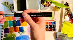 My hand holding the DaVinci Casaneo travel flat watercolor brush with colorful painting swatches in the background 