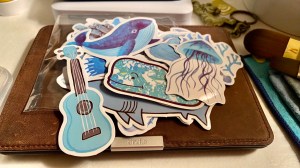 Many watercolor stickers laying on top of my kindle leather case on my desk