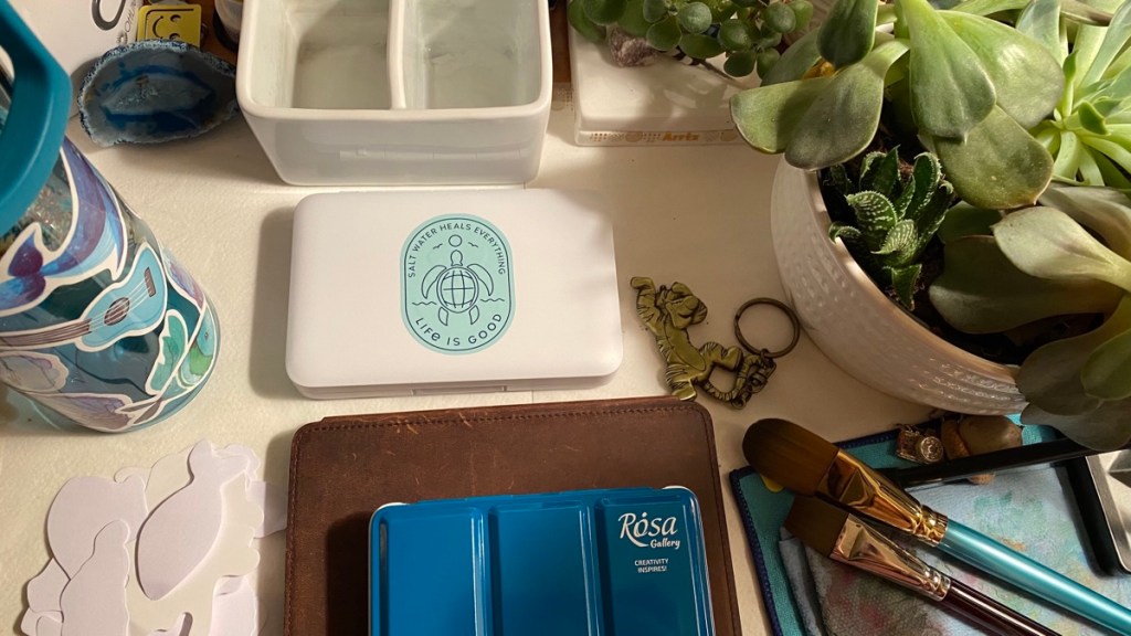 The sticker reads: salt water heals everything. Life is good. Sticker on Van Gogh watercolor pocket box on my desk. 