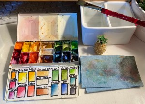 Medium whiskey painters palette on my desk with full pans and half pans of watercolor in it. To the right is a water container with a brush, a cloth, and a pineapple keychain. 